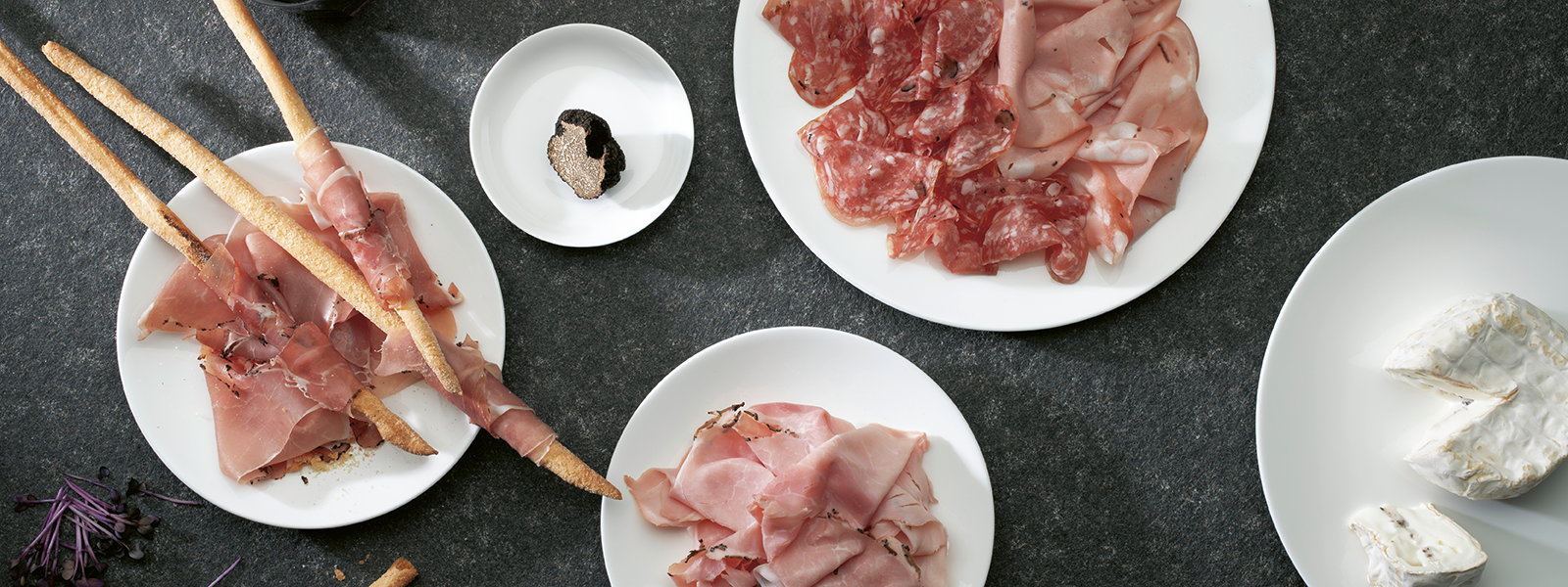 Ein Bild mit vier Tellern, darauf angerichtet sind: Trüffel Salami, Trüffelschinken, ein Trüffelkäse, eine Trüffelknolle und Grissinis, die mit Schinken umwickelt sind.